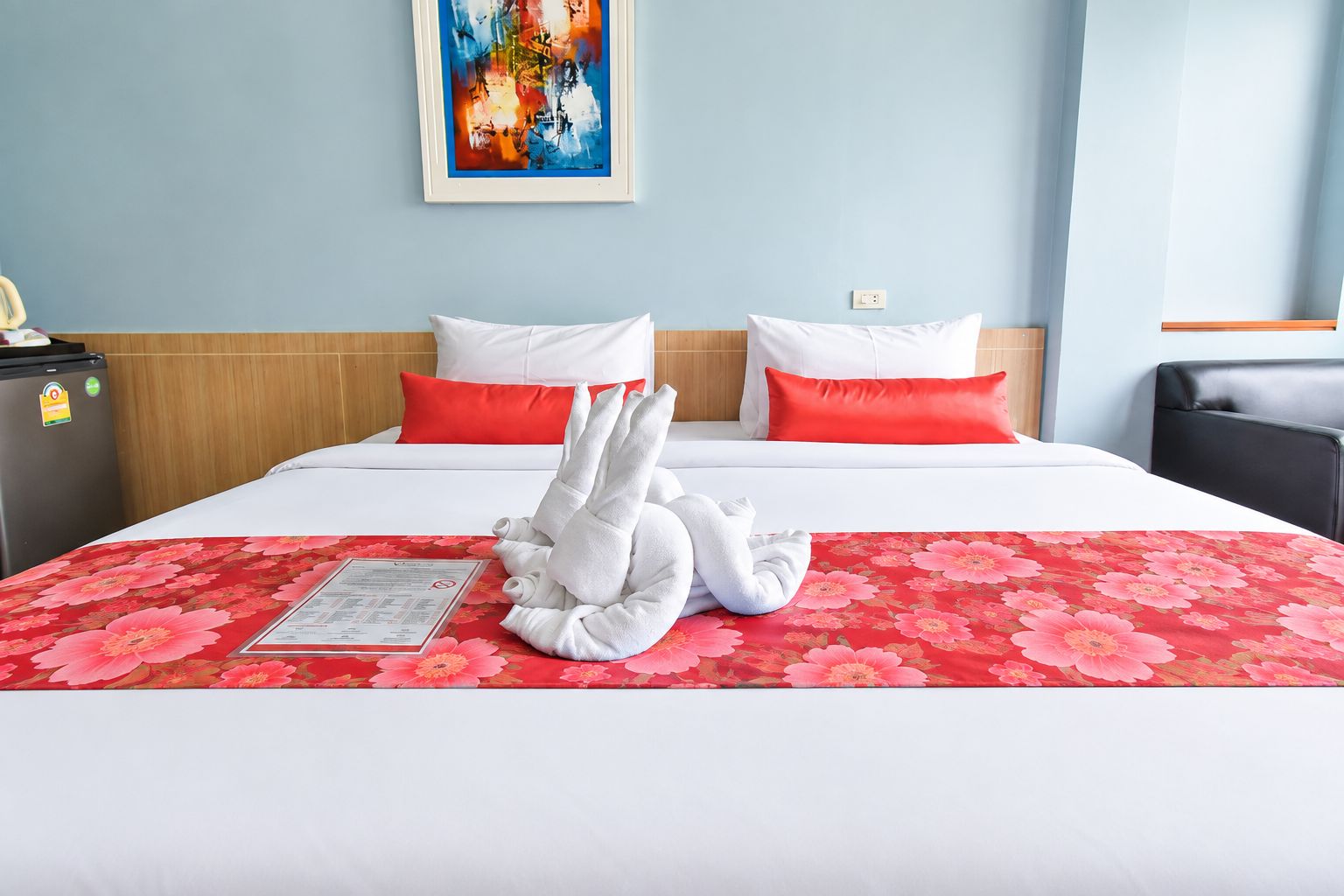 Habitación de hotel con una cama, dos almohadas, una colcha roja con estampado floral, una escultura de toalla blanca, un cuadro en la pared y un escritorio con refrigerador.