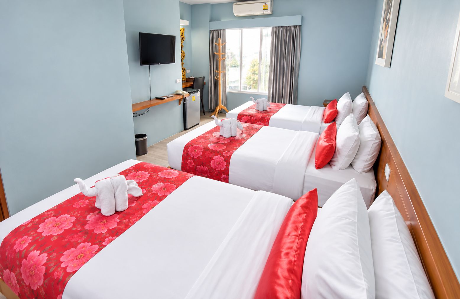 Habitación de hotel con tres camas, cada una con cubrecamas floreados de color rojo y almohadas blancas. Hay un televisor montado en la pared, un escritorio con una silla y una ventana con cortinas.