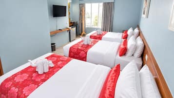 Habitación de hotel con tres camas, cada una con cubrecamas floreados de color rojo y almohadas blancas. Hay un televisor montado en la pared, un escritorio con una silla y una ventana con cortinas.
