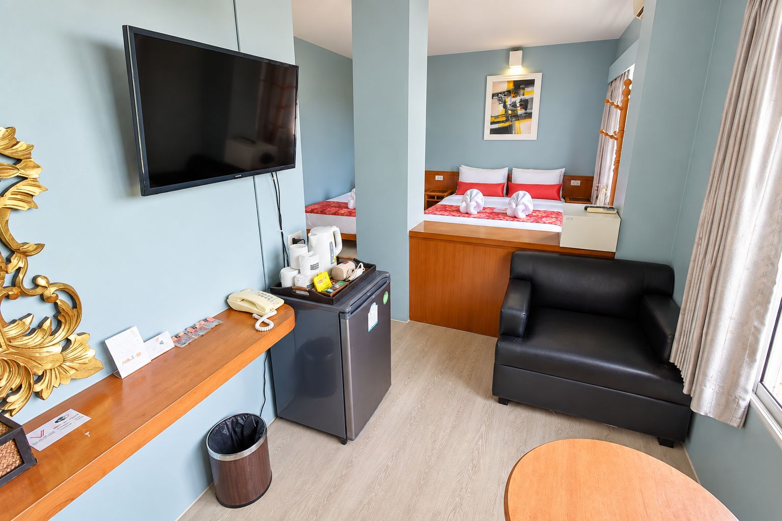 Habitación de hotel con televisión de pantalla plana, escritorio con teléfono, cubo de basura, sofá de cuero negro, una mesita redonda y cama con colcha roja y blanca.