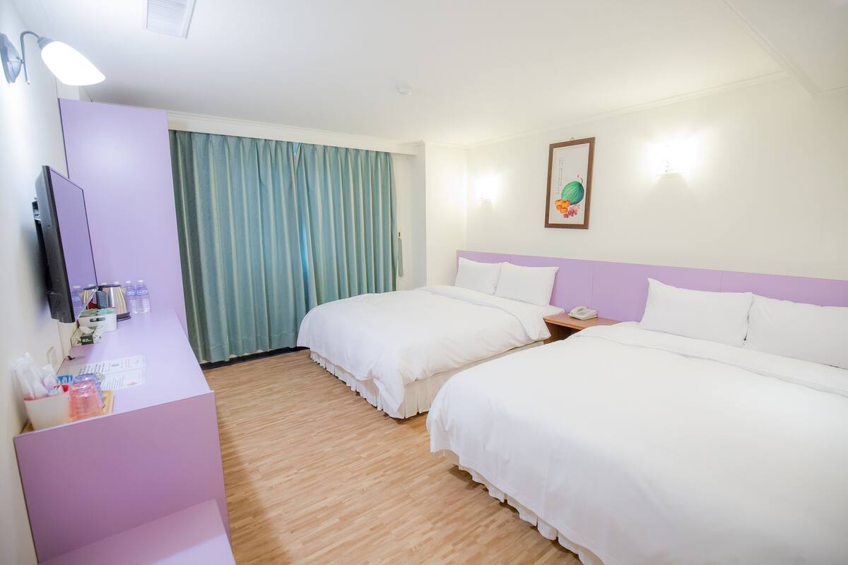 A hotel room with two beds, a desk with a computer, a telephone, and a window with curtains.