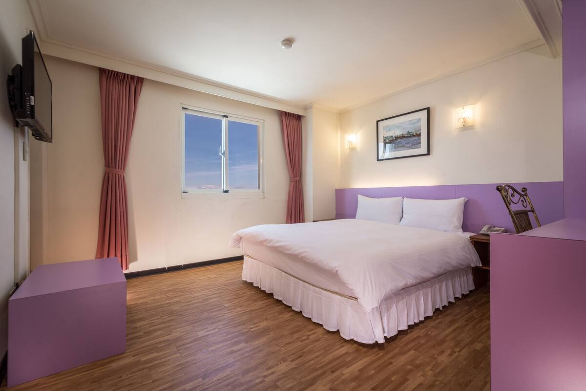 A bedroom with a large bed, a window, a flat-screen TV, and a bedside table.