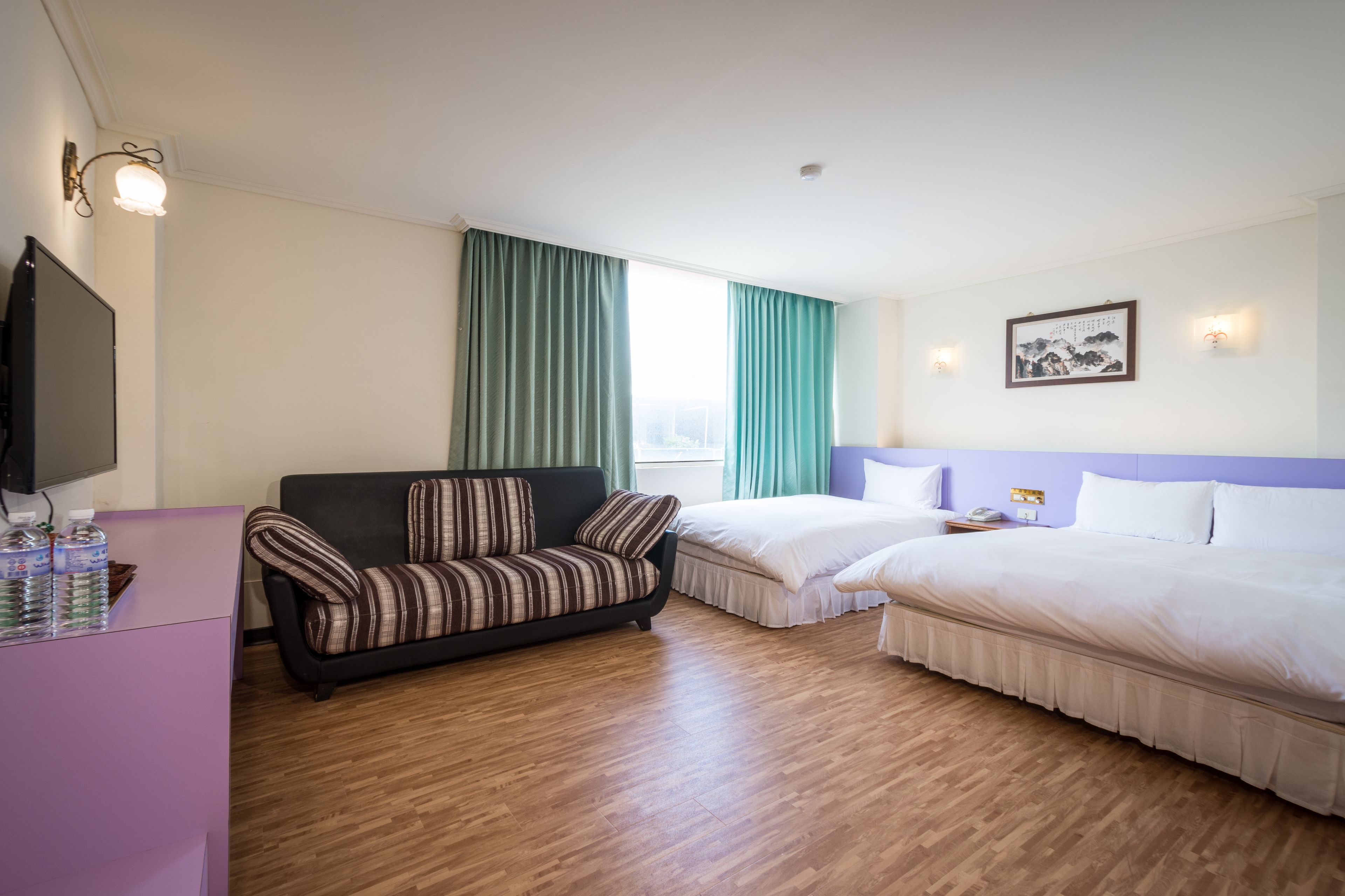 A hotel room with a sofa, a bed, a desk with a TV, and a window with curtains.