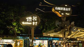 A night scene with illuminated signs in a public area, possibly a park or market, with people and buildings in the background.