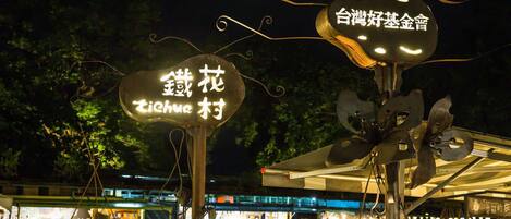 A night scene with illuminated signs in a public area, possibly a park or market, with people and buildings in the background.