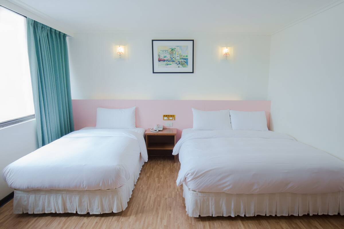 A hotel room with two single beds, a wooden floor, a window with curtains, a nightstand with a phone, and a wall-mounted painting.