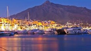 A marina with boats docked, a mountain in the background, and buildings with lights.