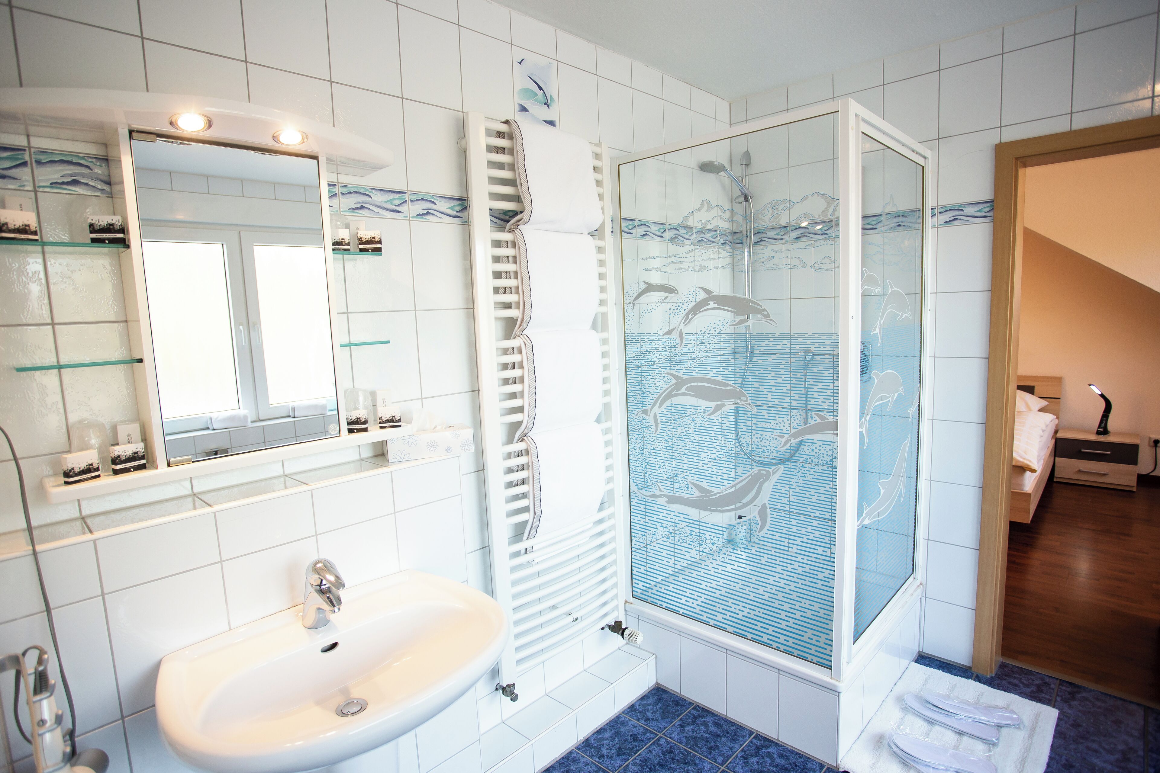 Een badkamer met een witte wastafel, een douchecabine met glazen wanden en een afbeelding van dolfijnen, en een betegelde vloer.