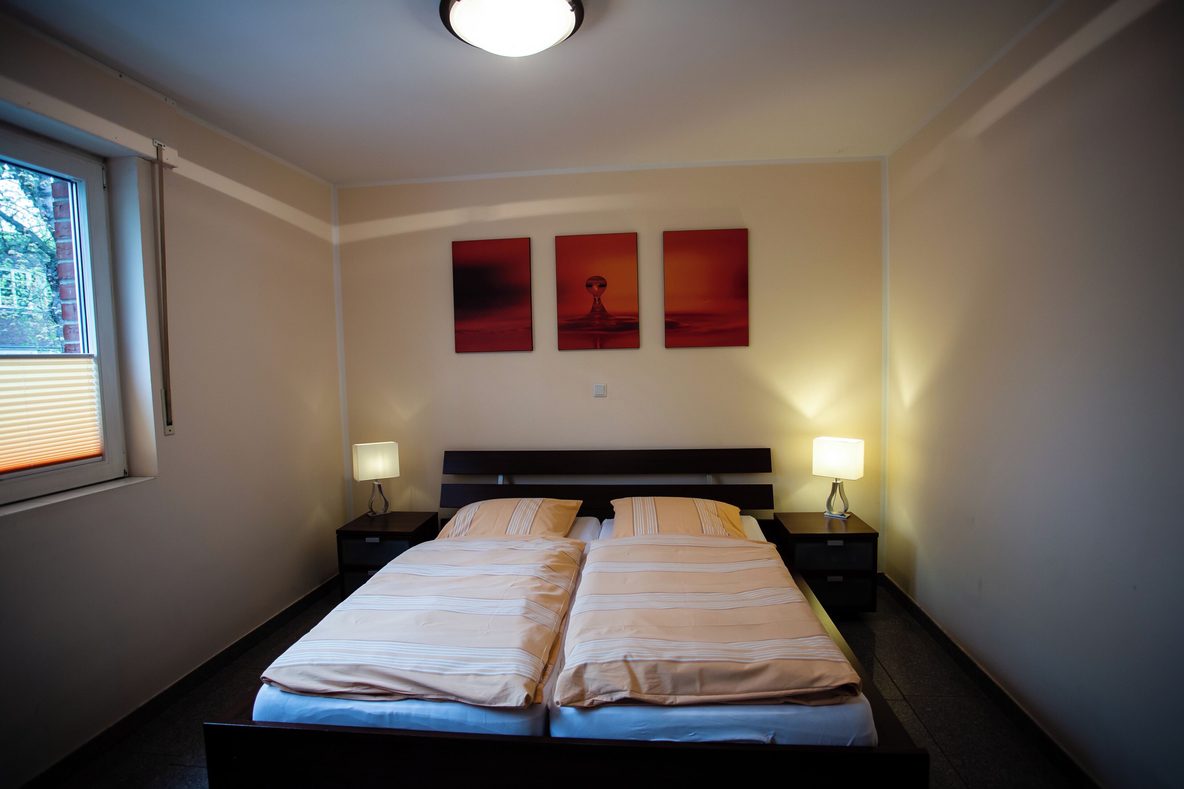 Una cama doble con dos mesitas de noche, cada una con una lámpara, en una habitación con una ventana y tres obras de arte colgadas en la pared.