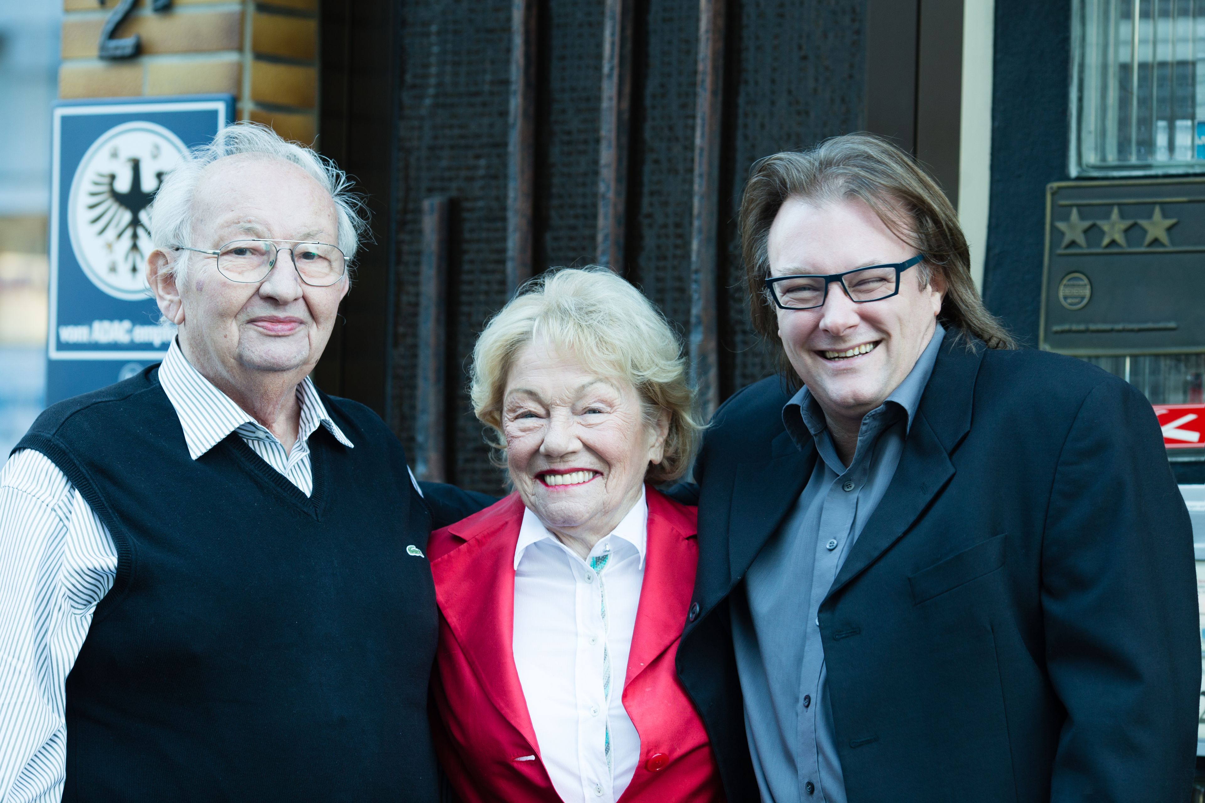 Drie personen staan voor een gebouw met een bord en een plaquette.