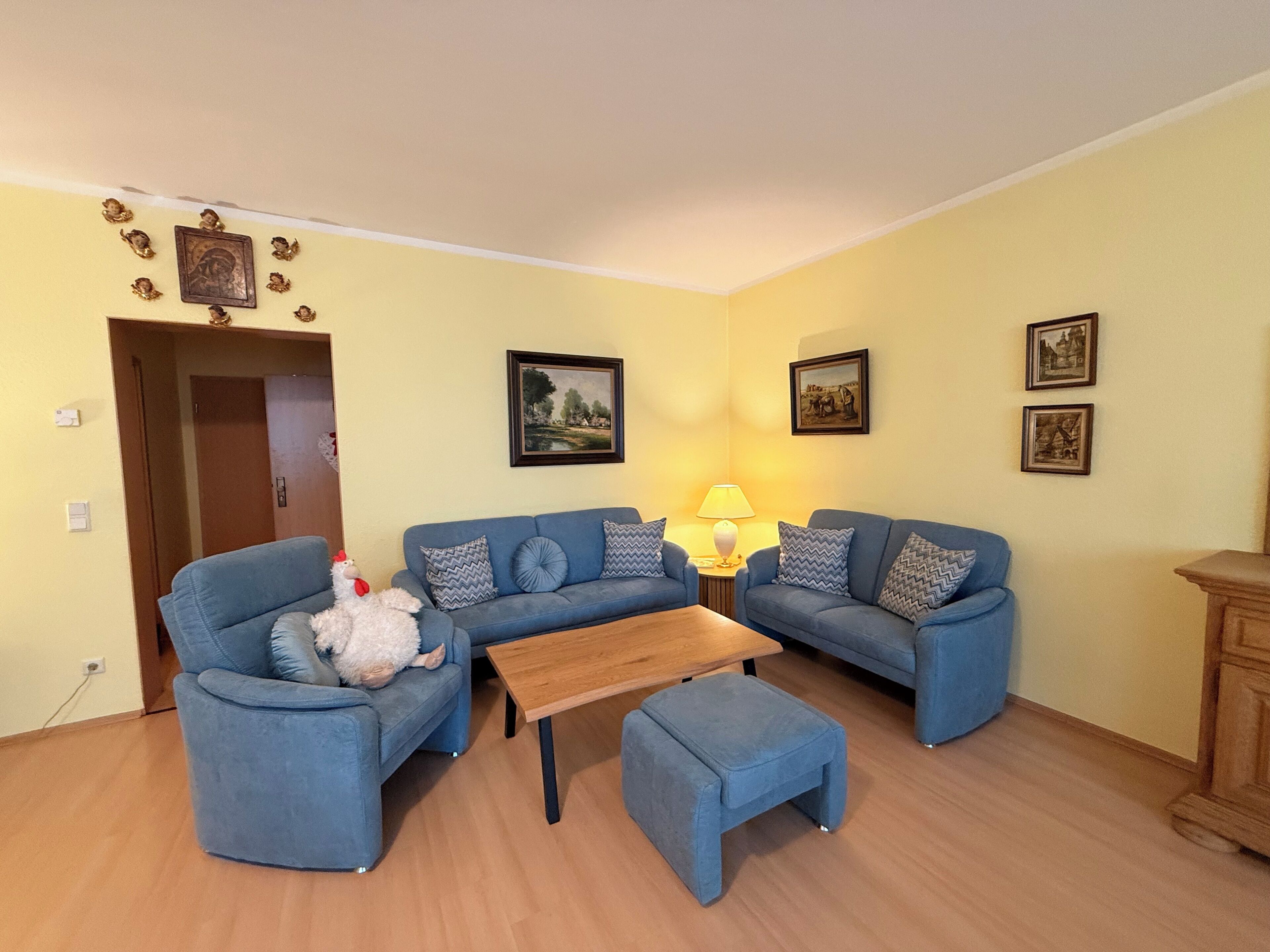 Una sala de estar con dos sofás azules, una mesa de centro de madera y un pequeño otomaní. Hay cuadros enmarcados en las paredes y una lámpara sobre la mesa de centro.