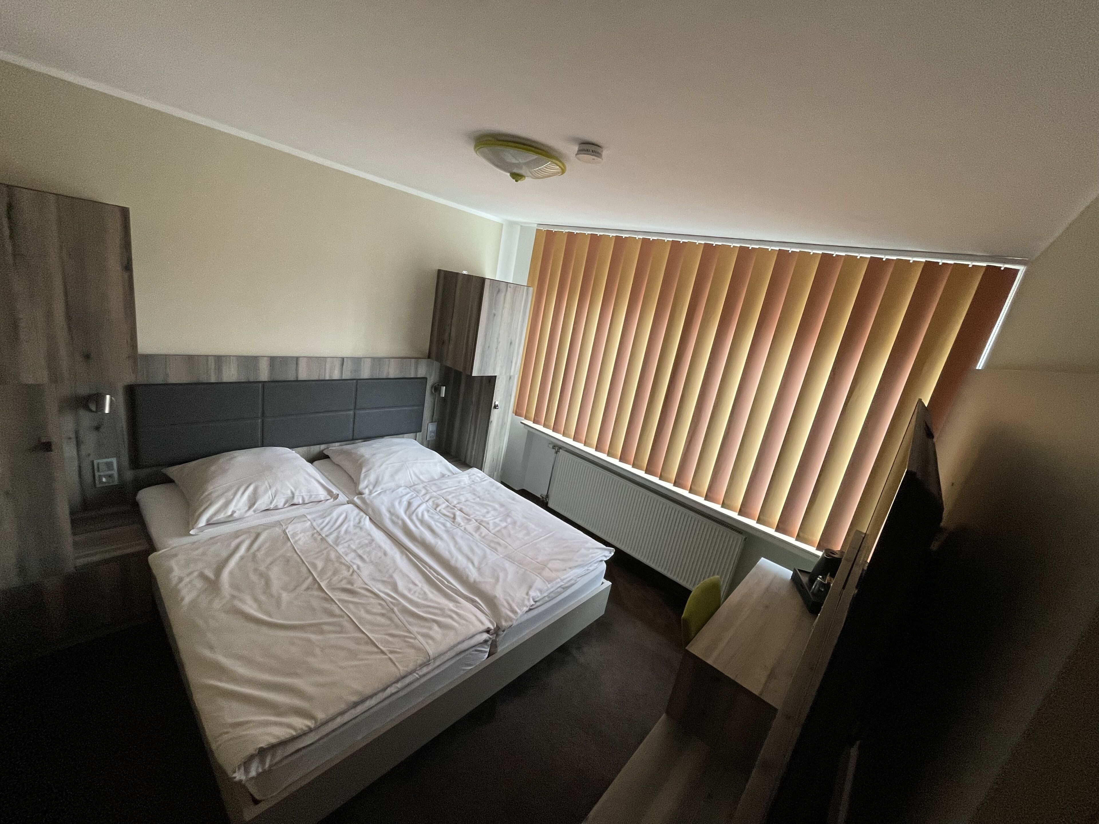 Un dormitorio con cama, cabecero de madera, mesita de noche, lámpara y ventana con persianas.