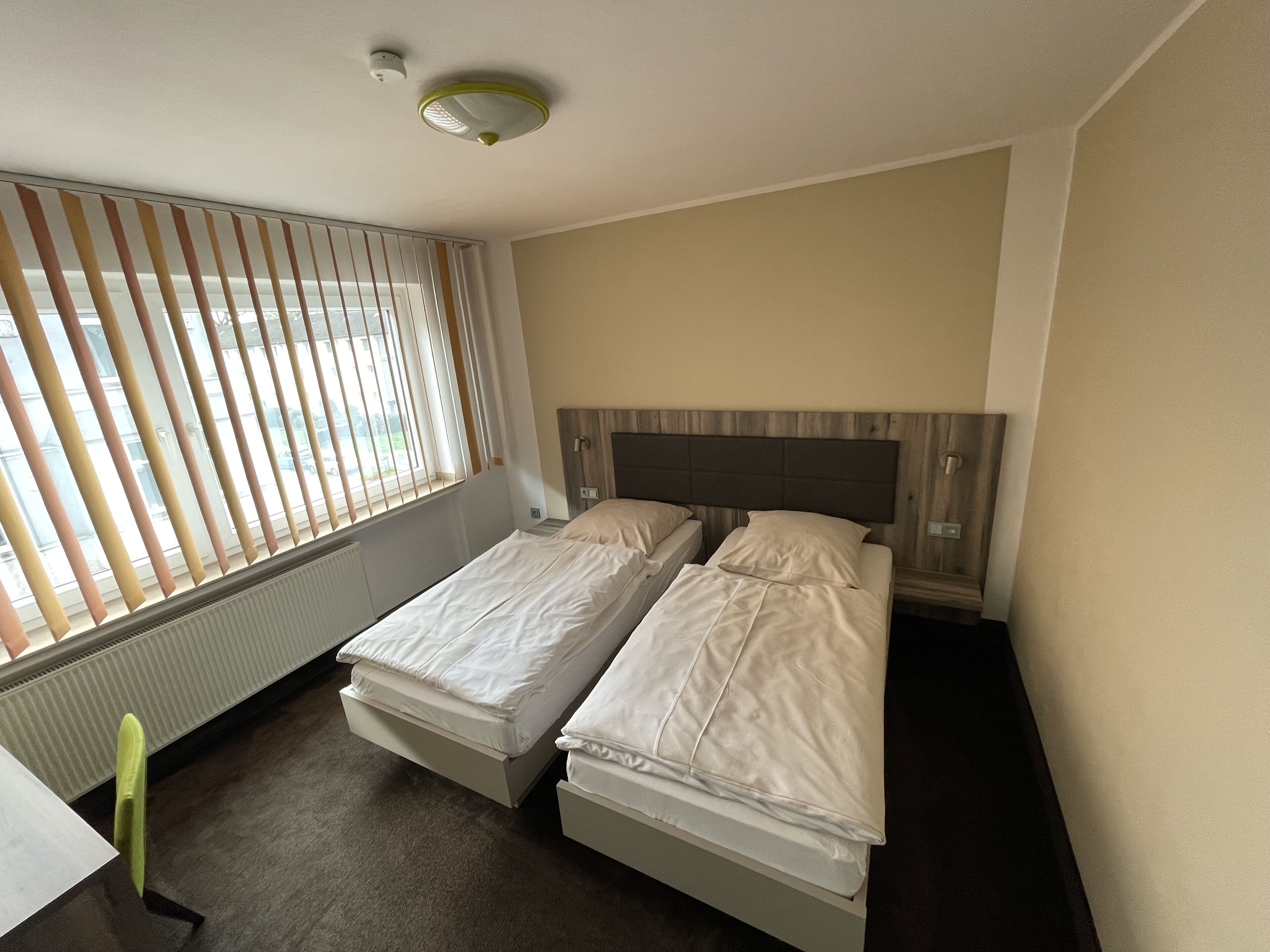 Een hotelkamer met twee éénpersoonsbedden, een radiator, een raam met zonwering en een plafondlamp.