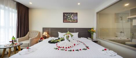 A hotel room with a neatly made bed, a sitting area with a chair and a small table, a bedside lamp, a painting on the wall, and a bathroom with a shower and toilet.