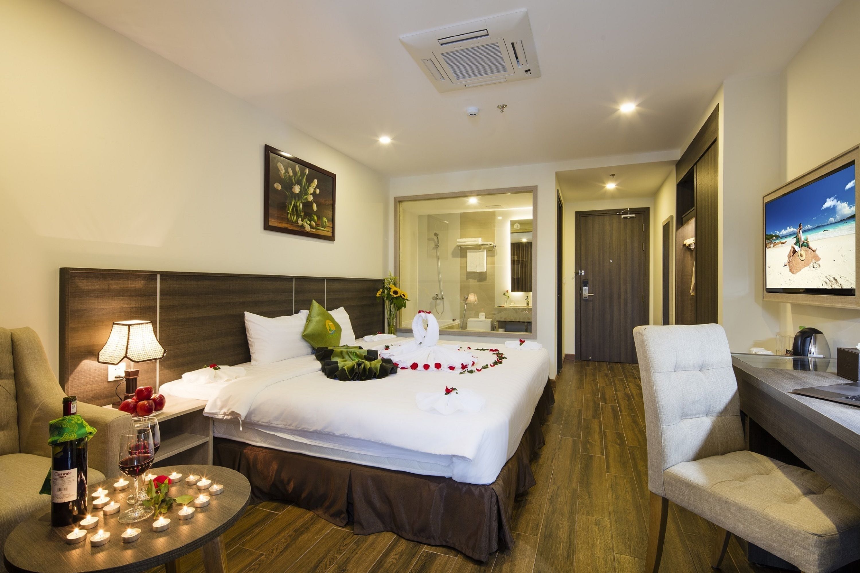 Quarto de hotel com cama bem arrumada, escrivaninha com cadeira, luminária e garrafa de vinho.