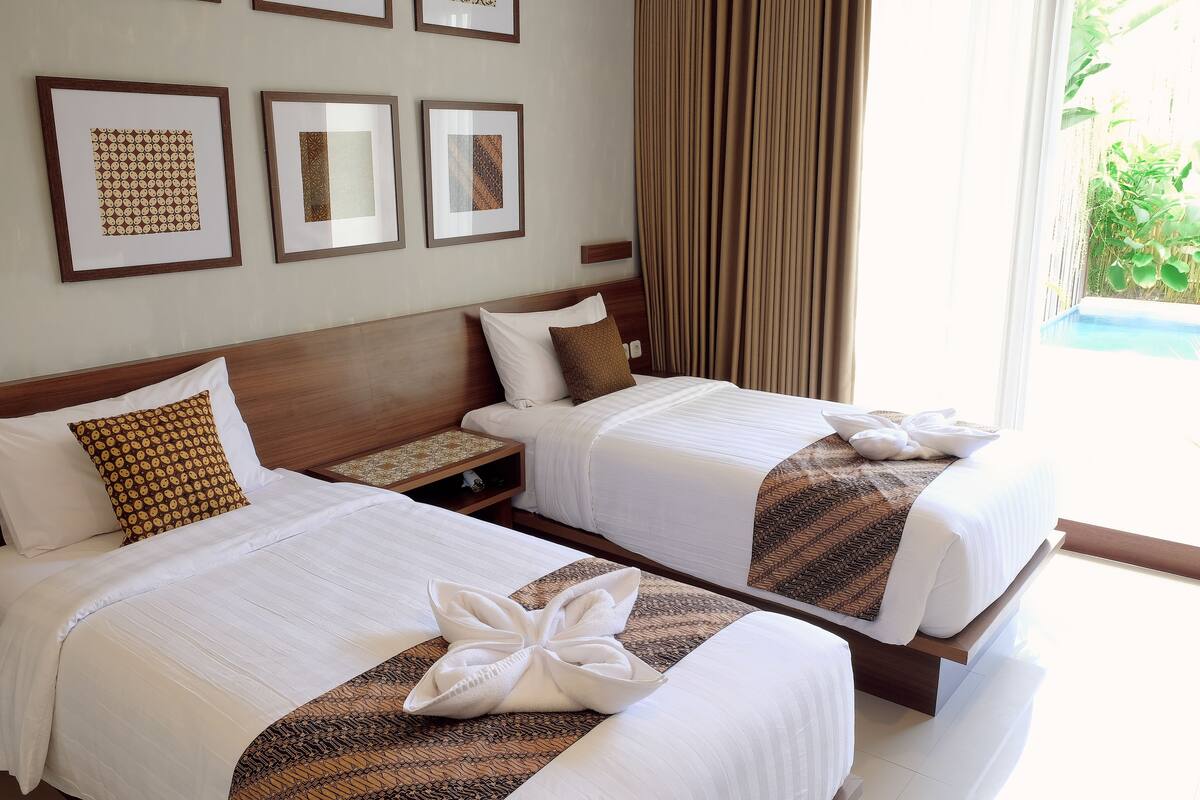 A hotel room with two beds, each with a white bedspread and a brown patterned pillow. There are framed artworks on the wall and a window with curtains.
