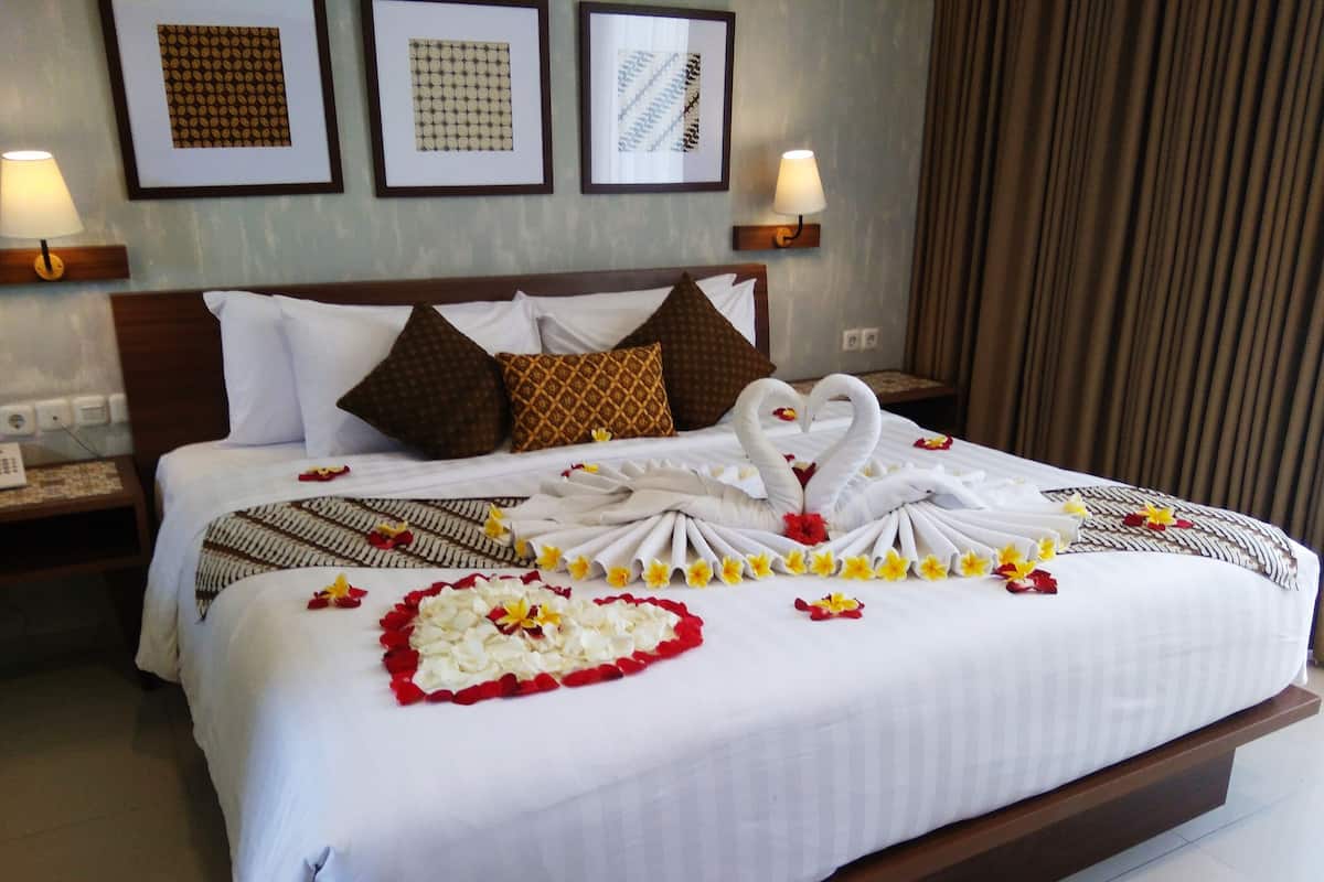 A neatly made bed with a heart-shaped arrangement of towels and flowers, a wooden headboard, and a wall with framed pictures.