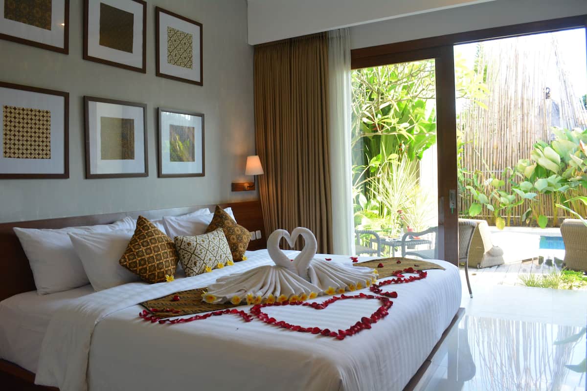 A hotel room with a bed, framed pictures on the wall, a window with curtains, and a view of a pool and greenery outside.