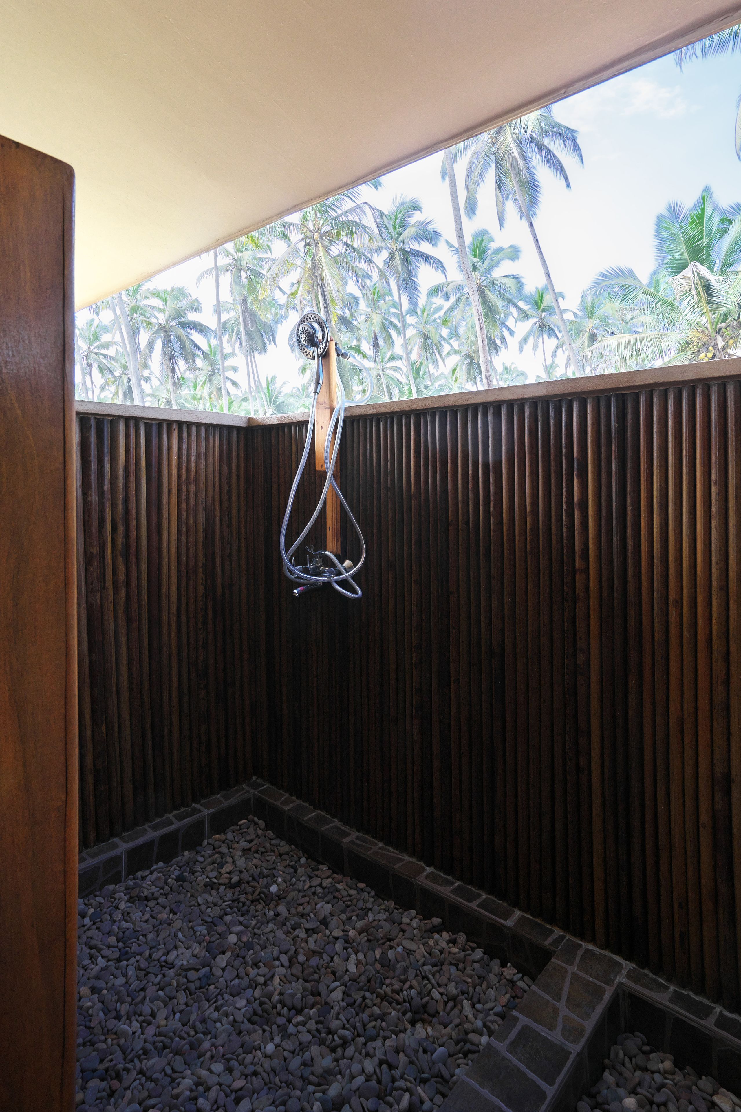 A wooden-framed shower with a stone floor, a hanging showerhead, and a view of palm trees through a window.