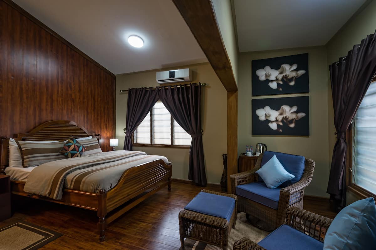 A bedroom with a wooden bed, a wicker chair, a blue ottoman, and two orchid paintings on the wall.