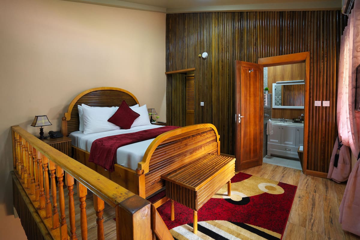 A bedroom with a wooden bed, a wooden bench, a red and beige rug, a wooden wardrobe, and a bathroom visible through an open door.