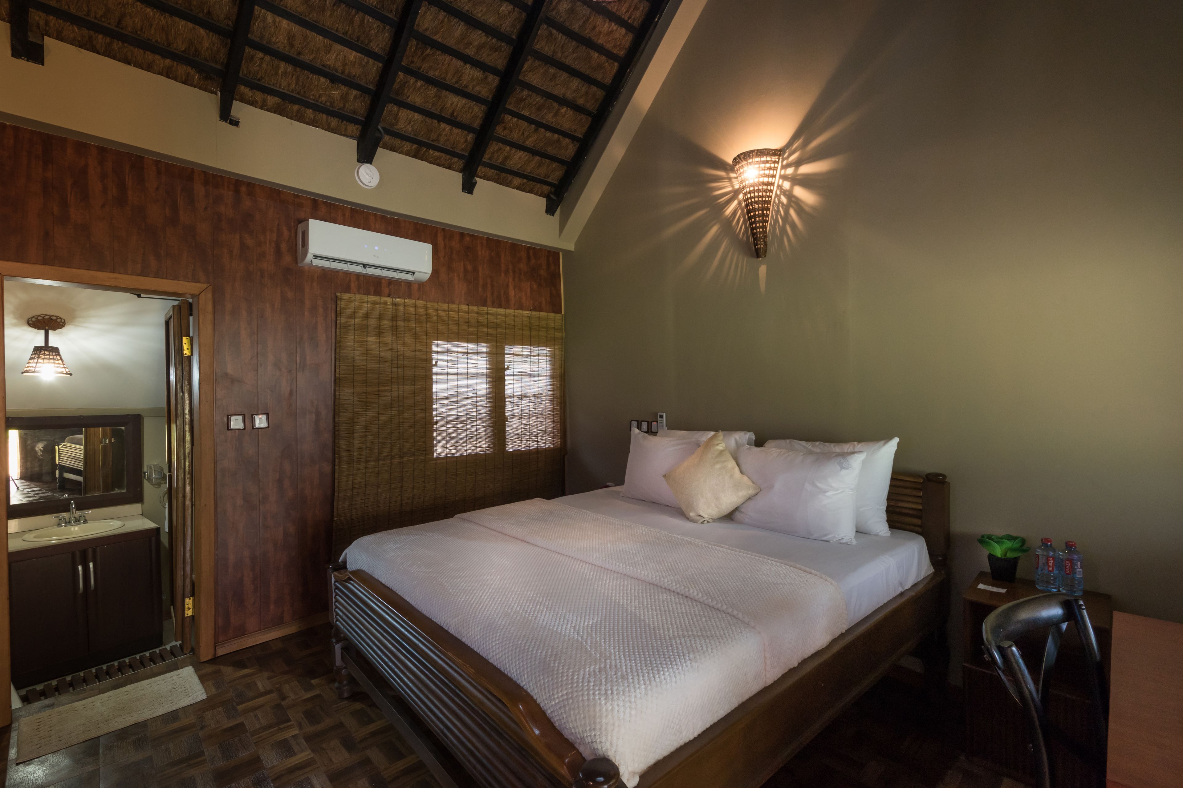 A bedroom with a wooden bed, a nightstand with a lamp, a window with blinds, and a ceiling fan.