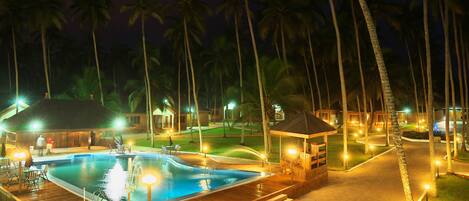 Un área de piscina nocturna con cubierta de madera, palmeras y estructuras iluminadas.