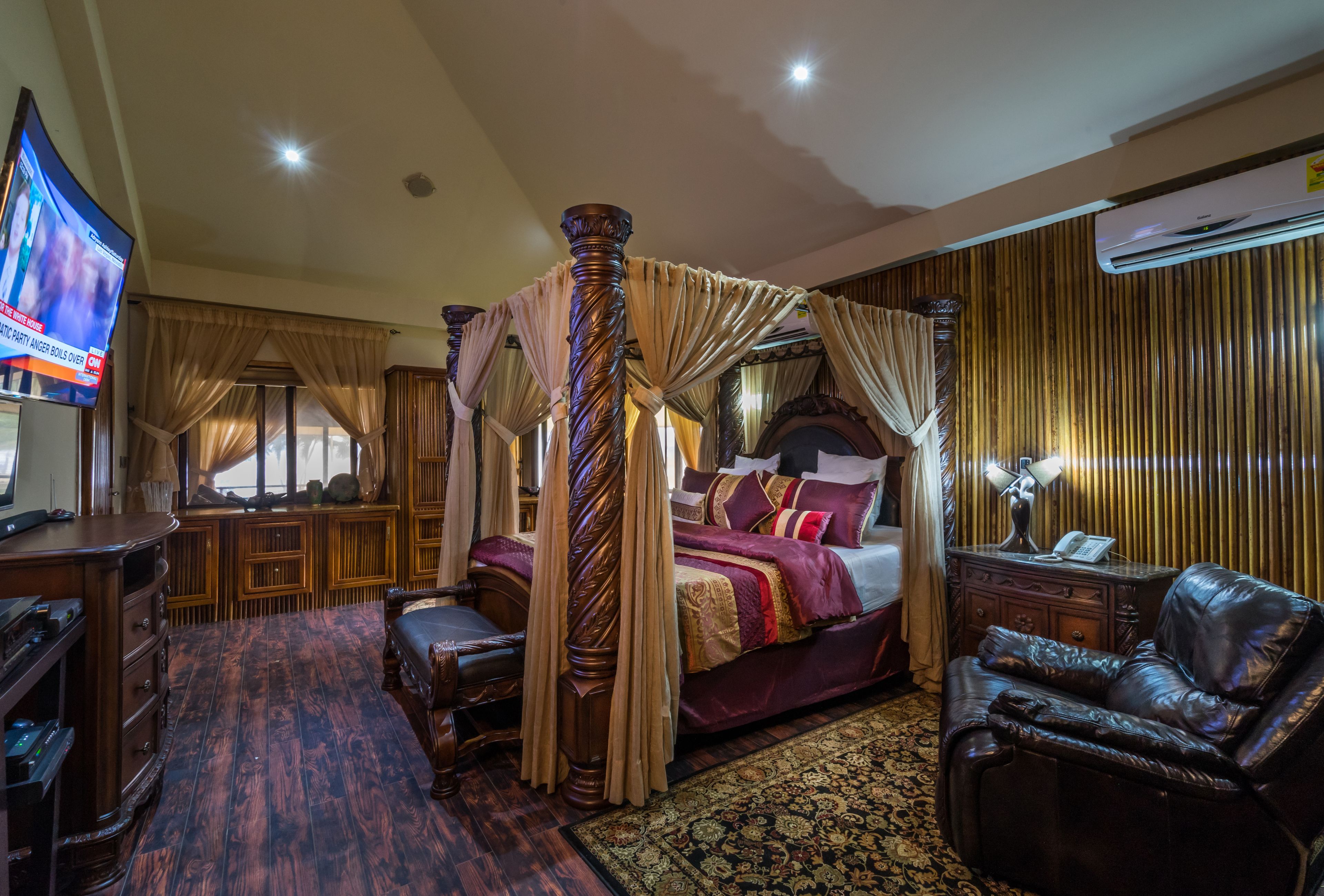 A luxurious bedroom with a four-poster bed, a leather armchair, a television, and a decorative rug.