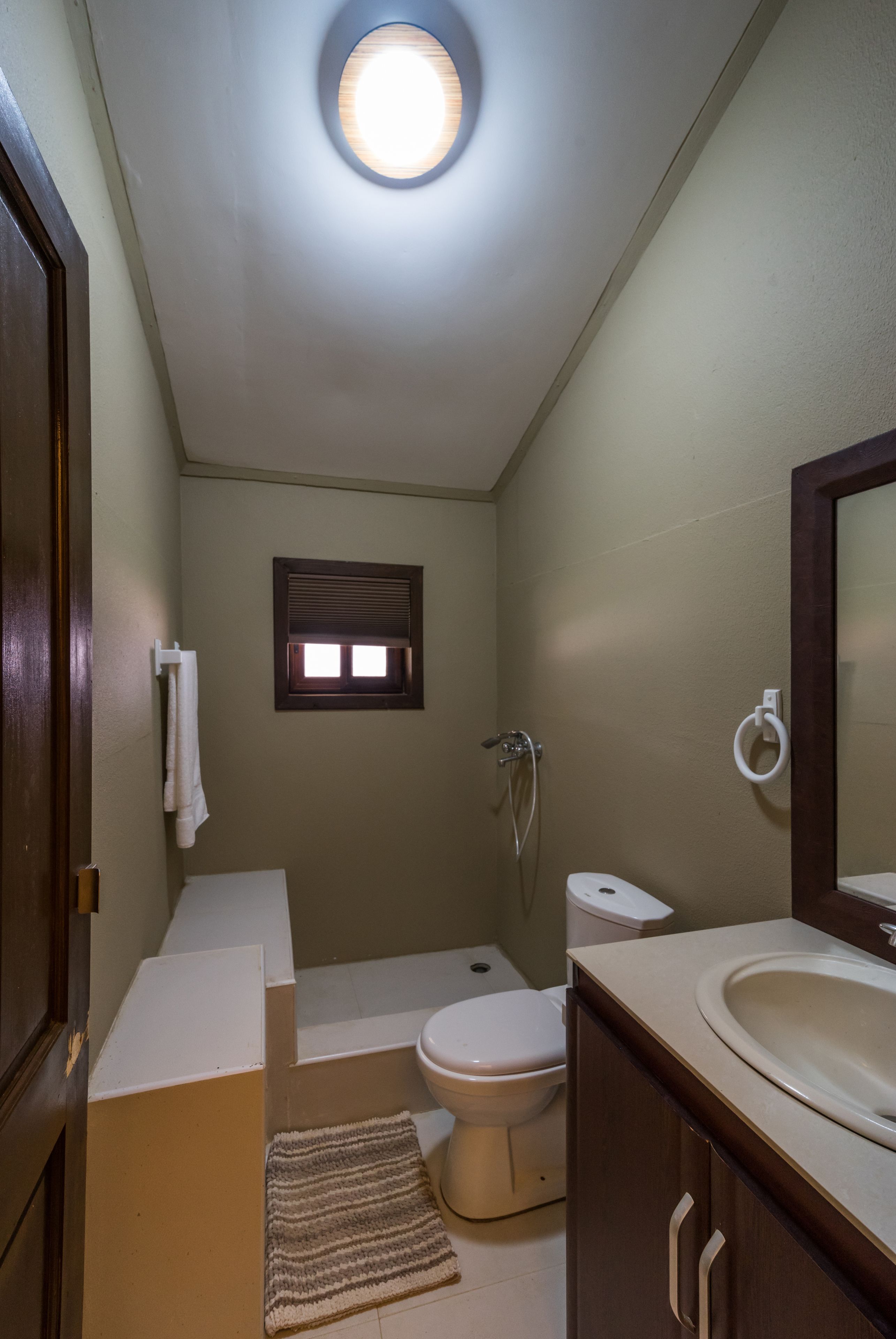 A small bathroom with a toilet, sink, and shower. There is a window above the toilet and a towel hanging on the wall.