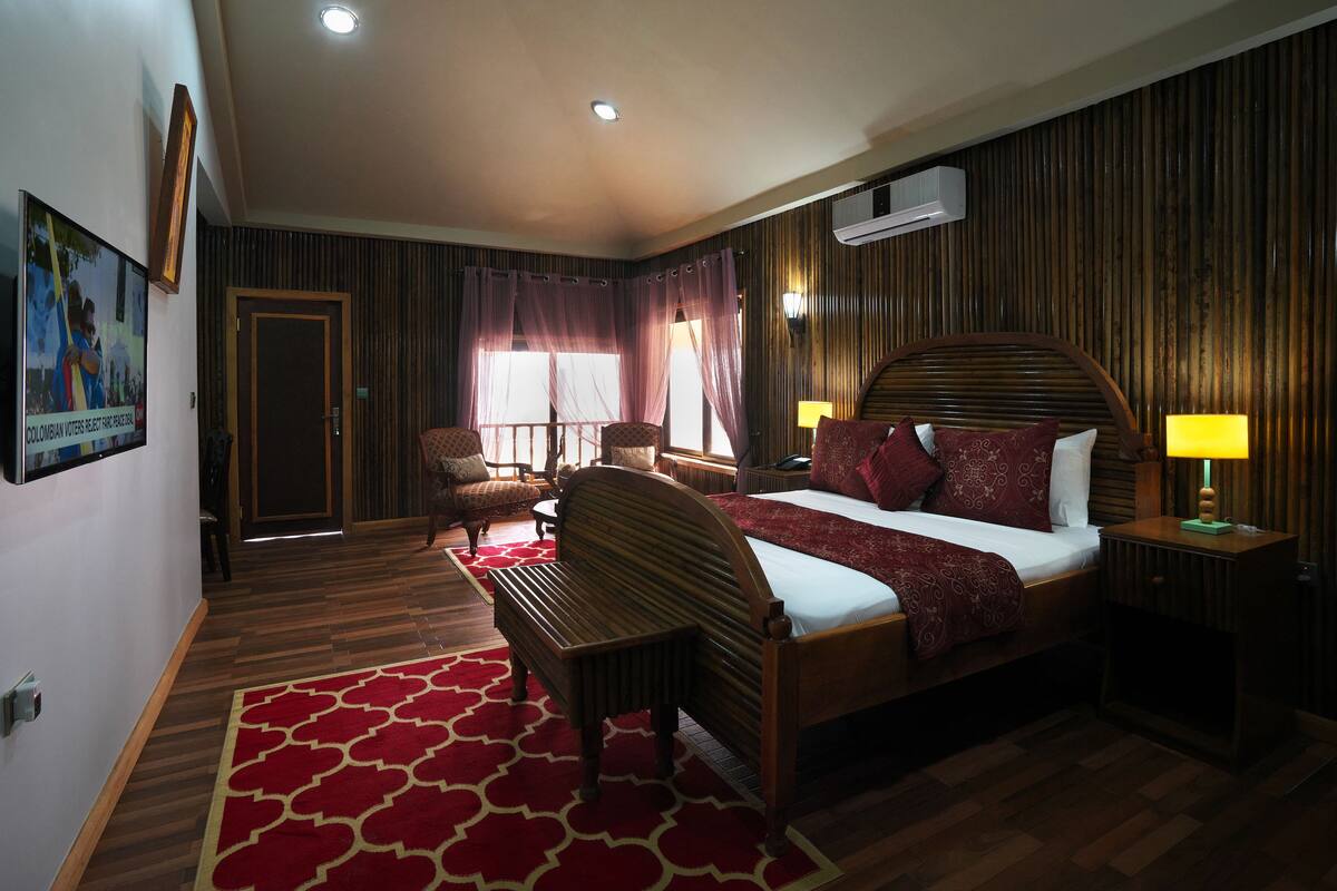 A hotel room with a large bed, a wooden headboard, a bench, a desk, and a chair. There is a red patterned rug on the floor, and the room has wooden paneling on the walls.