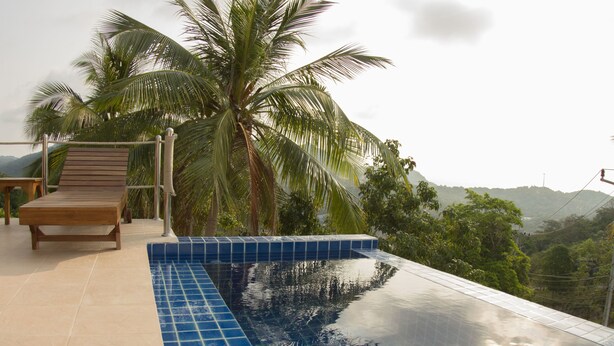 A pool with a clear blue tile edge, a wooden lounge chair, and a palm tree in the background. - Karo Villas (Koh Tao)