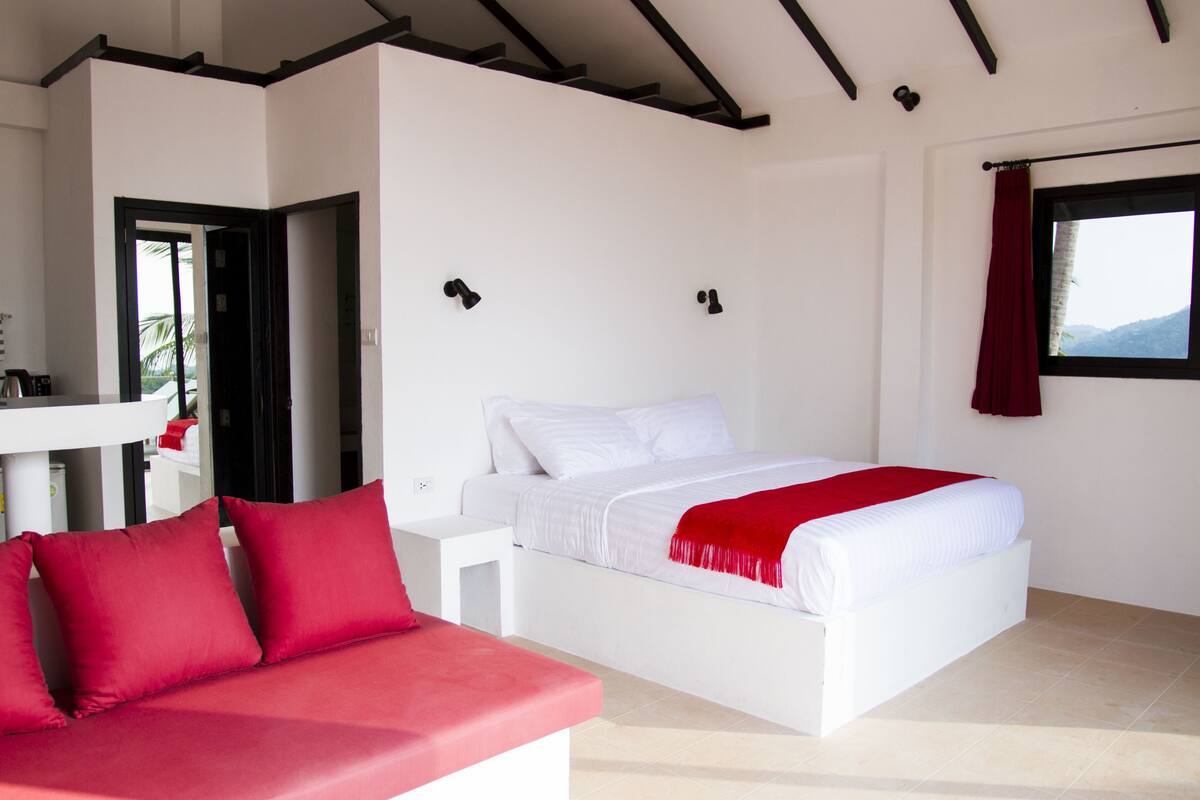 Ein Schlafzimmer mit einem weißen Bett, roten Kissen, einem weißen Nachttisch und einem roten Vorhang.