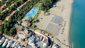 Eine Vogelperspektive zeigt einen Küstenort mit einer Marina, einem Schwimmbad und einem Strand mit Sonnenschirmen.