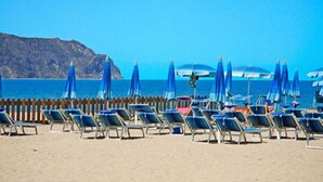 Ein Strand mit Reihen von Liegestühlen und blauen Sonnenschirmen, eine Holzumzäunung und ein klarer, blauer Himmel.