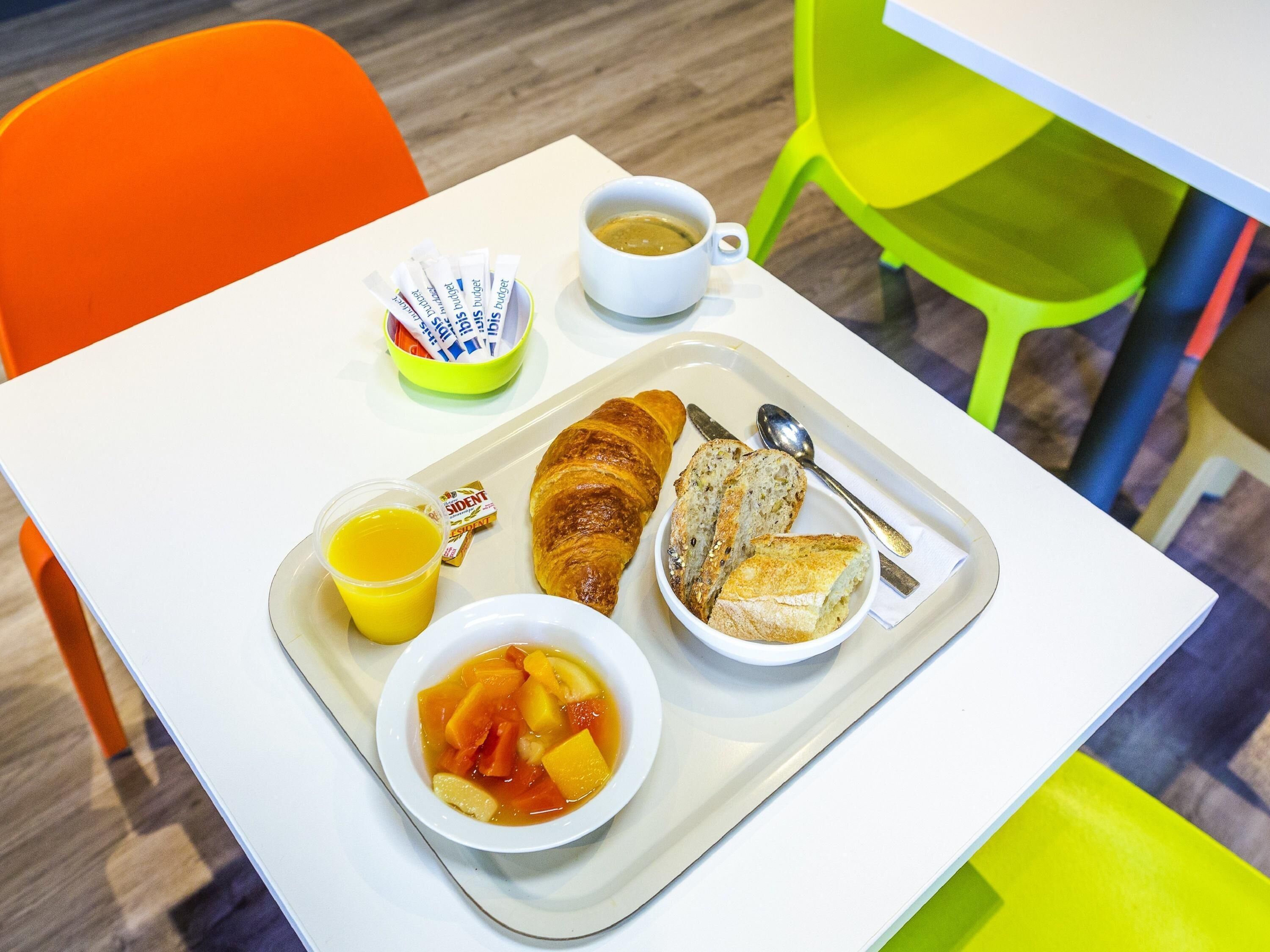 Una bandeja con cruasanes, pan, jugo de naranja y un tazón de sopa de frutas sobre una mesa blanca.
