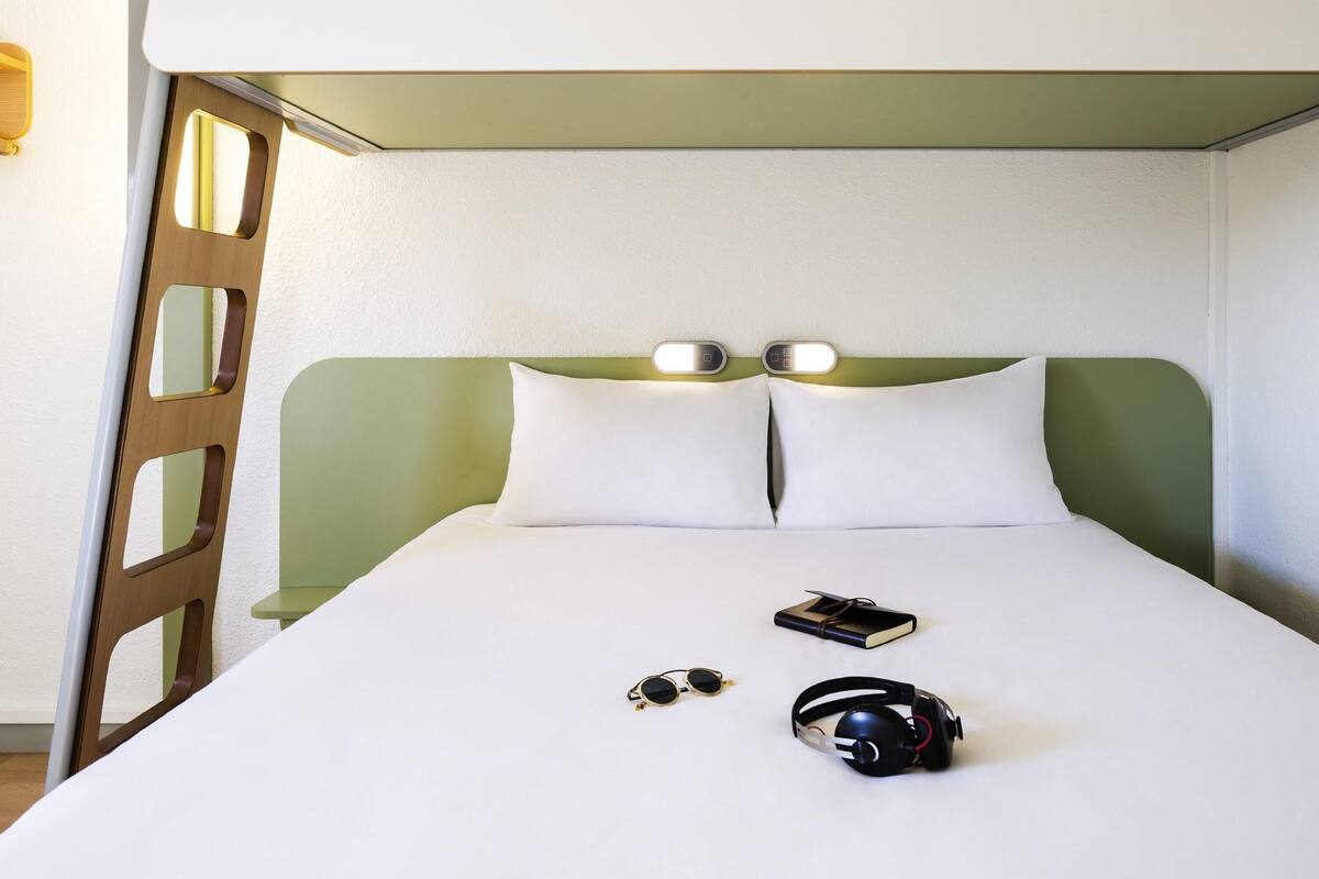 Een net opgemaakt bed met witte lakens, twee kussens, een boek, een paar zonnebrillen en een headset.