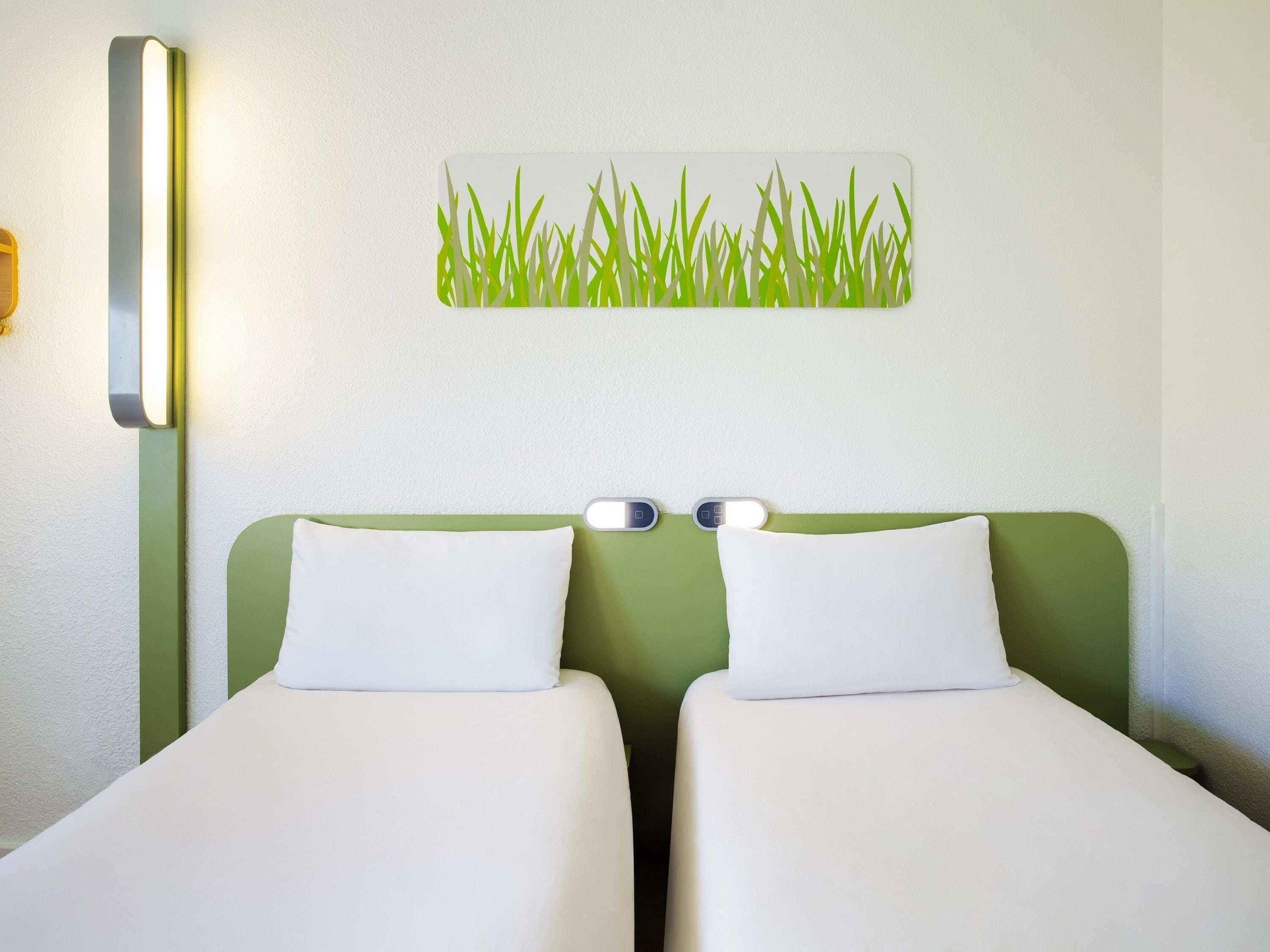 Ein Hotelzimmer mit zwei Einzelbetten, einem grünen Kopfteil, einer Wandlampe und einem Wandbild mit Gras.