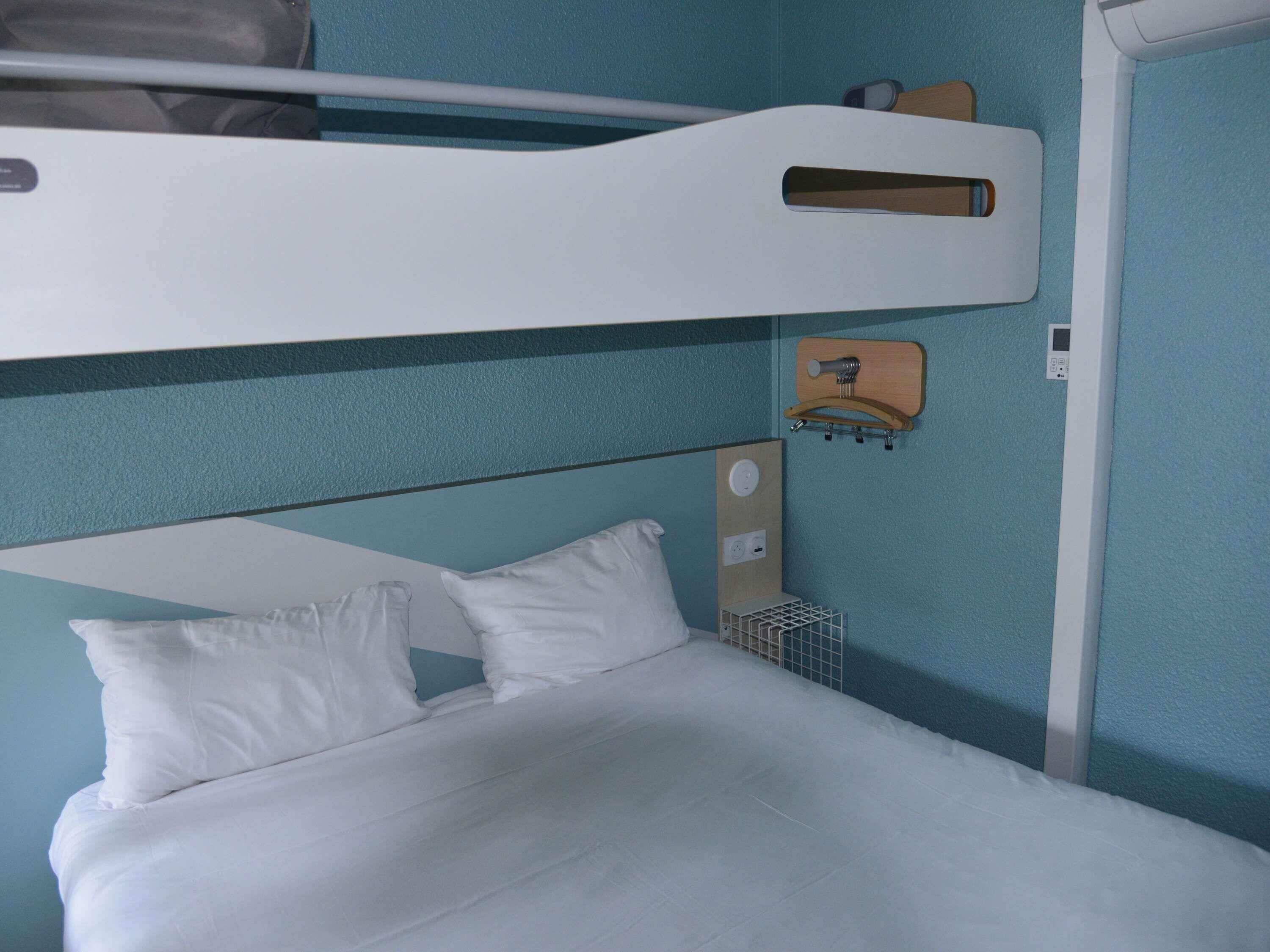 A bunk bed with a white mattress and pillows, a wooden headboard, a small wooden shelf, and a wall-mounted light fixture.