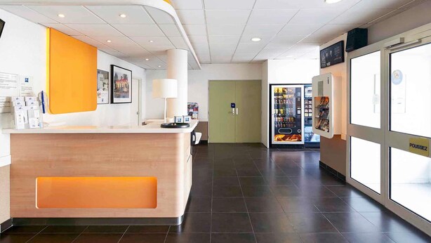 A reception area with a wooden reception desk, a vending machine, and a door in the background. - ibis budget Blois Centre (Blois)