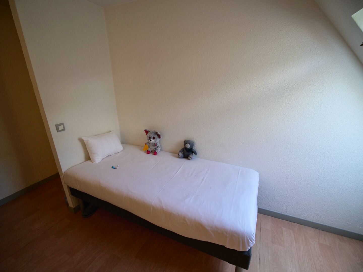 Una habitación pequeña con cama individual, colchón blanco y suelo de madera. Hay dos peluches en la cama.