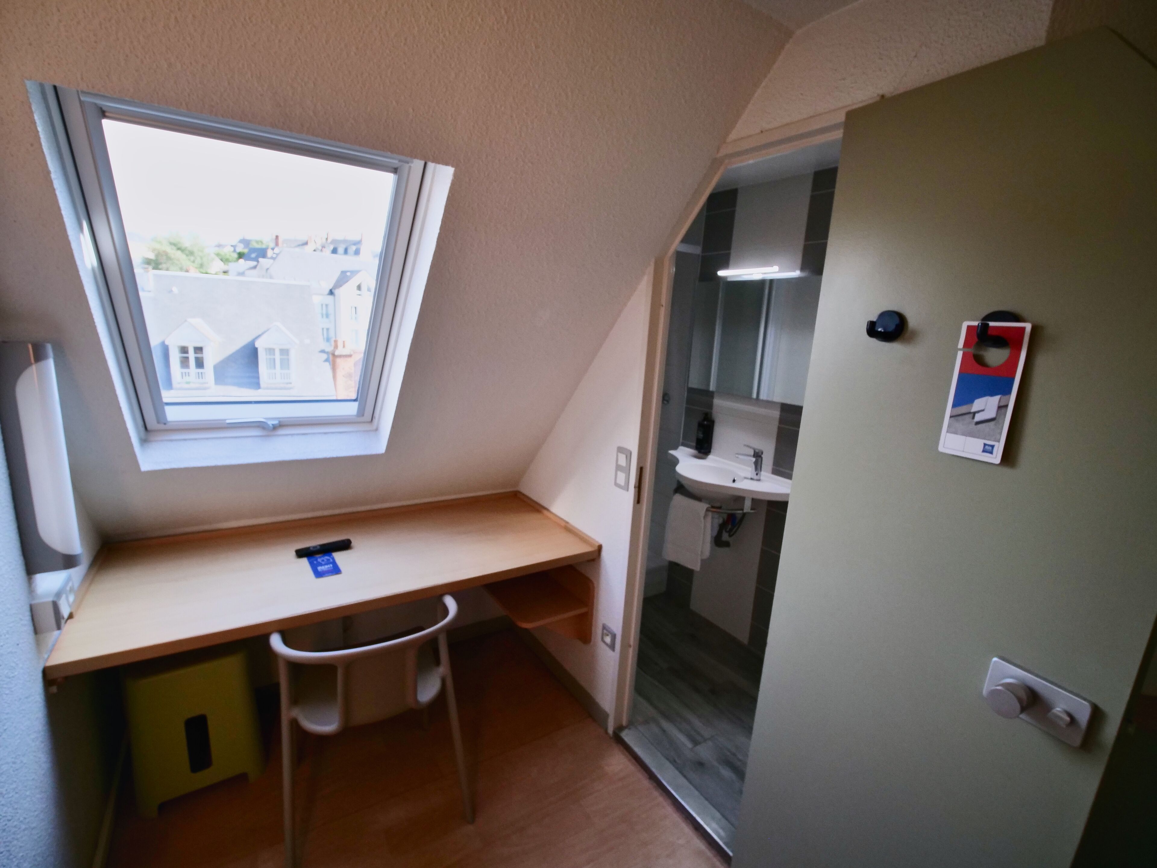 A small room with a sloped ceiling, a window, a desk, a chair, a sink, and a door.