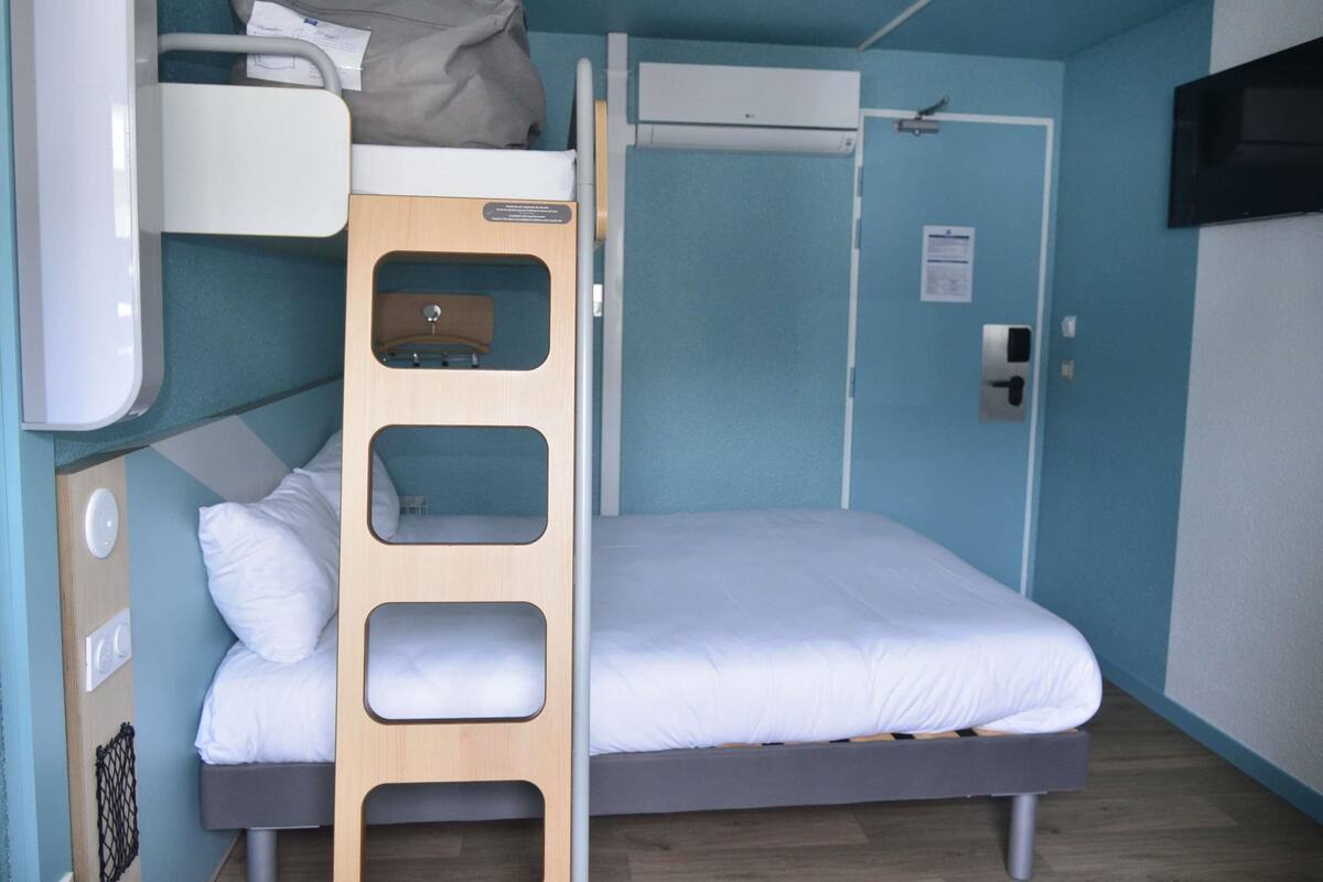 Une pièce de taille modeste, équipée d’un lit superposé, d’un petit bureau, d’un téléviseur fixé au mur et d’une porte dotée d’une poignée.