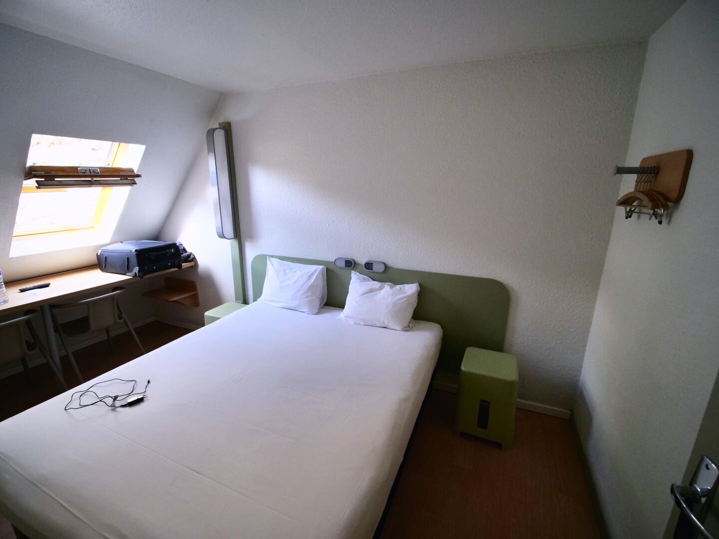 Un dormitorio con cama, escritorio, silla, taburete pequeño, ventana y estantería en la pared.