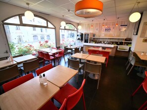 A restaurant with tables and chairs, a kitchen area, and large windows with a view of buildings and flowers outside. - ibis budget Blois Centre (Blois)