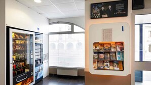 A vending machine with snacks and drinks, a bulletin board with travel brochures, and a view of a building through a window. - ibis budget Blois Centre (Blois)