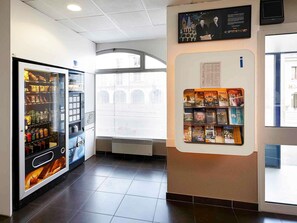 Una máquina expendedora con aperitivos y bebidas, un tablón de anuncios con folletos turísticos y una vista de un edificio a través de una ventana.