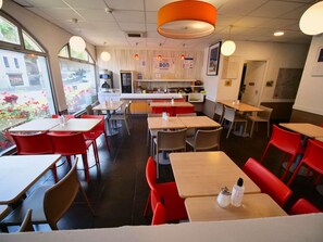 A restaurant interior with tables and chairs, a kitchen area, and a view through large windows. - ibis budget Blois Centre (Blois)