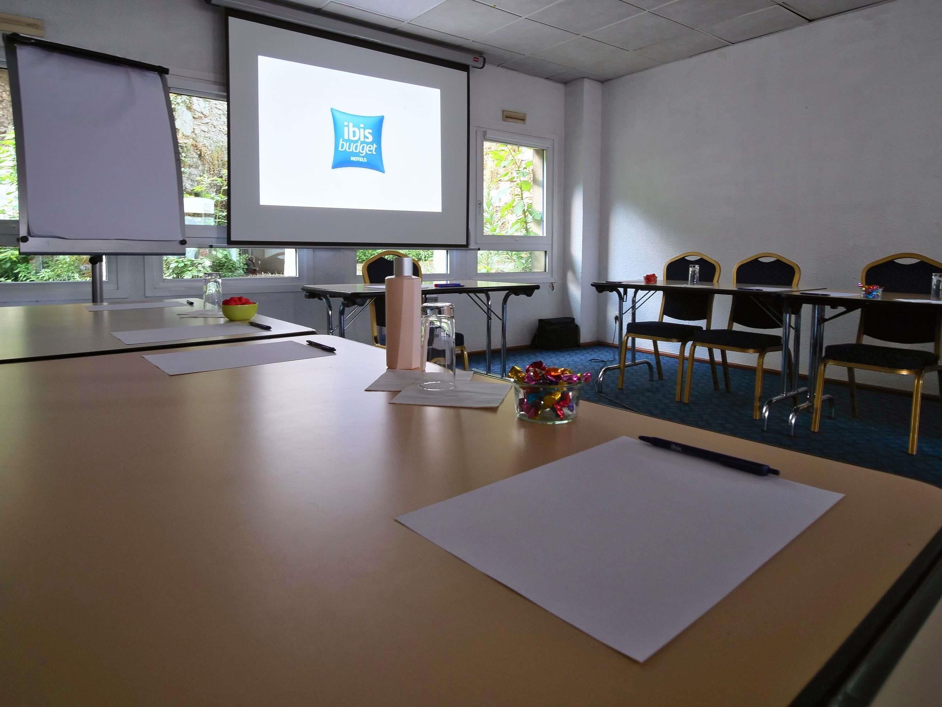 Una sala de conferencias con una pantalla de proyector mostrando el logotipo del hotel ibis budget, una pizarra, un jarrón de cristal con cintas de colores, y una mesa de madera con papeles y un bolígrafo.