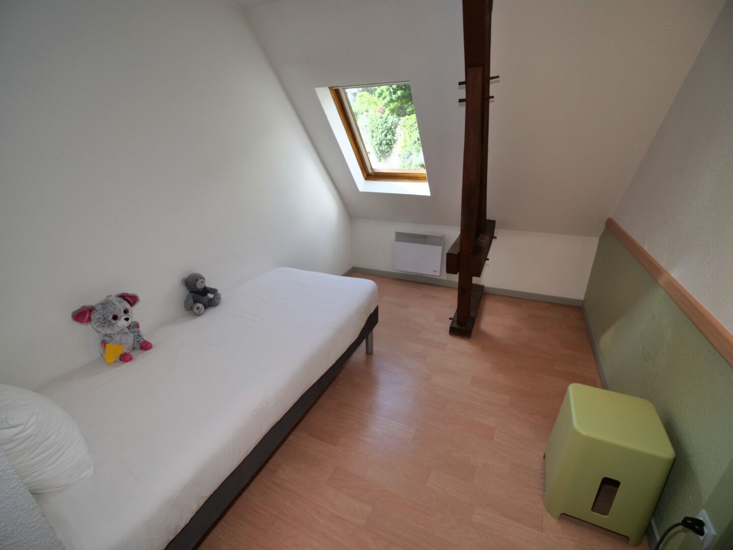 Een klein appartement met één kamer, een schuine plafond, dakraam, een bed, een klein krukje en een radiator.