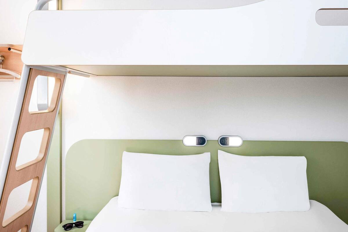 Un lit superposé avec une tête de lit verte, une literie blanche et une petite table de chevet sur laquelle se trouvent une bouteille et des lunettes de soleil.